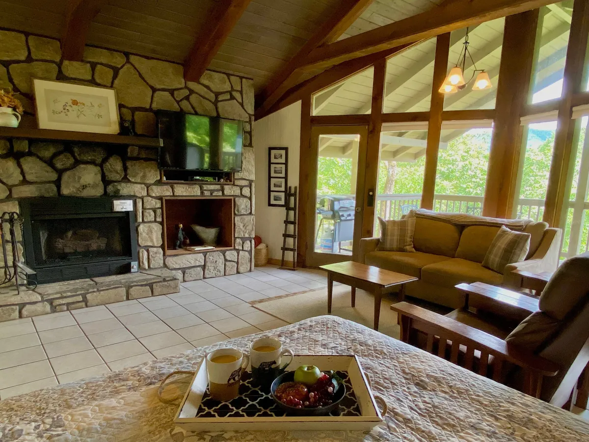 The image depicts a cozy living room with a prominent stone fireplace, large windows that allow natural light to flood the space, plush sofas and accent chairs arranged for conversation, and a coffee table with a tray. The warm, inviting atmosphere is enhanced by the soft lighting and view of the outdoors through the expansive windows.