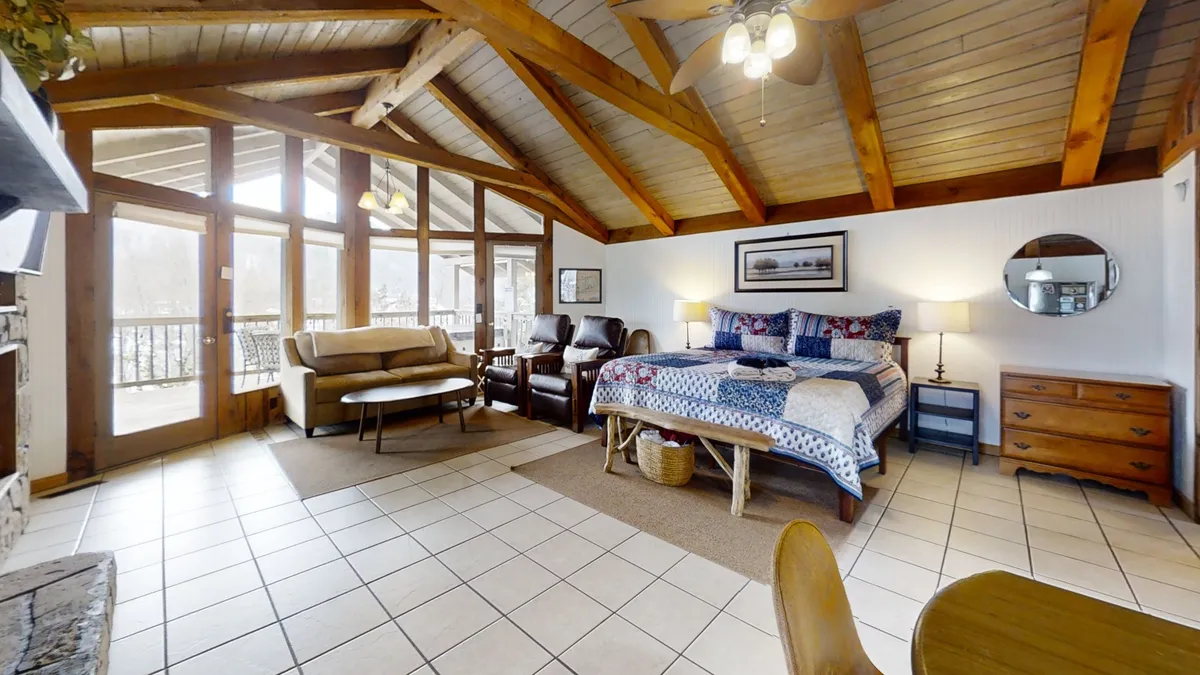 This image showcases an interior space with a large bed featuring patterned bedding, exposed wooden beams, large windows that fill the room with natural light, and a comfortable seating area. A small table near the window offers a cozy spot for breakfast or a casual meal, bathed in warm light.