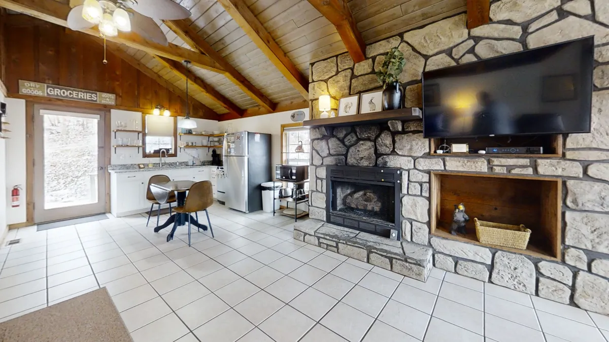 Showing an open-concept interior, this image features a stone fireplace, wooden beams, a visible kitchen, and a dining table. The space is designed for both relaxation and gatherings, with a warm, welcoming ambiance that encourages family activities and entertaining guests.