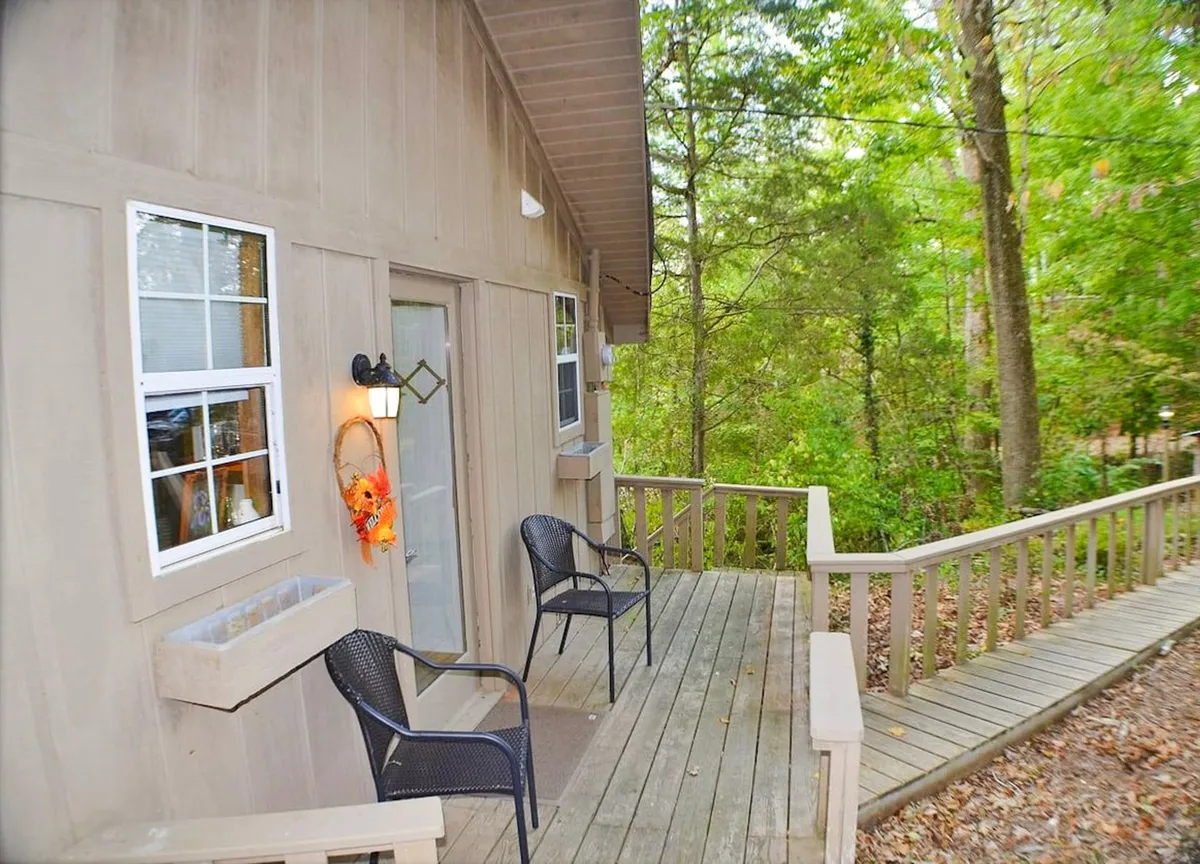 This image captures the exterior entrance of a cozy cabin, featuring a charming wooden pathway leading to the door. The sitting area with chairs encourages relaxing outdoors, and the decorative wreath adds a welcoming touch. The scene is surrounded by trees, providing a tranquil, nature-filled setting ideal for unwinding and enjoying the outdoors.