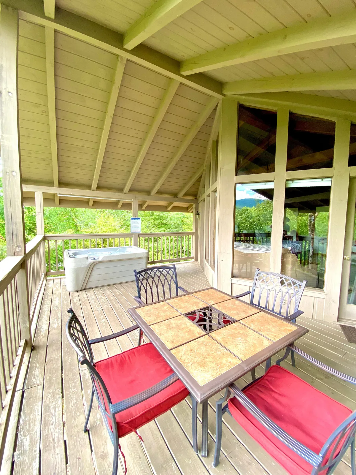 This image showcases a spacious wooden deck featuring a dining table with four chairs and a bubbling hot tub. The area is surrounded by lush greenery, inviting guests to relax and unwind. The deck is partially covered, providing shelter and comfort for outdoor dining or soaking in the hot tub while enjoying the natural setting.